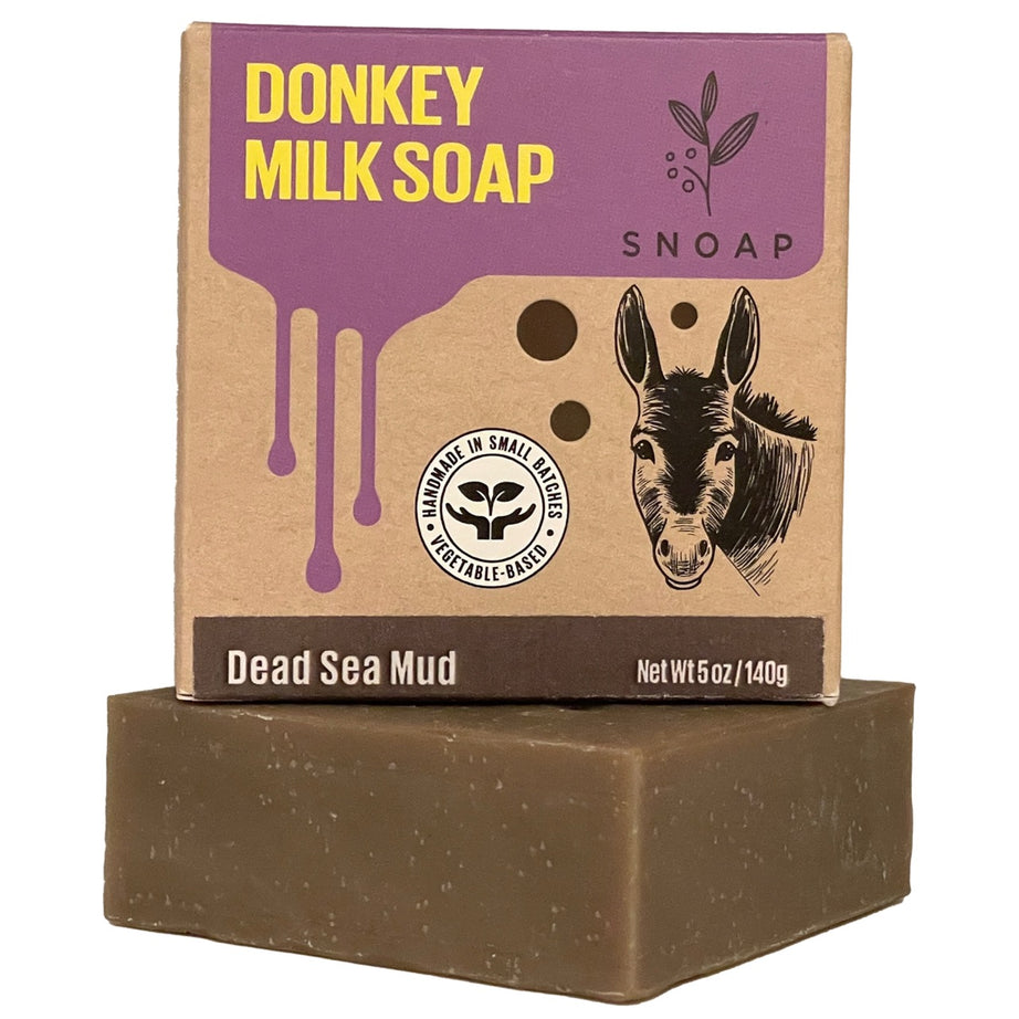 A bar of dead sea mud donkey milk soap with a dark brown color is stacked below its cardboard box packaging. The box is decorated with a sketch of a donkey and the text 'Donkey Milk Soap' and 'SNOAP'.