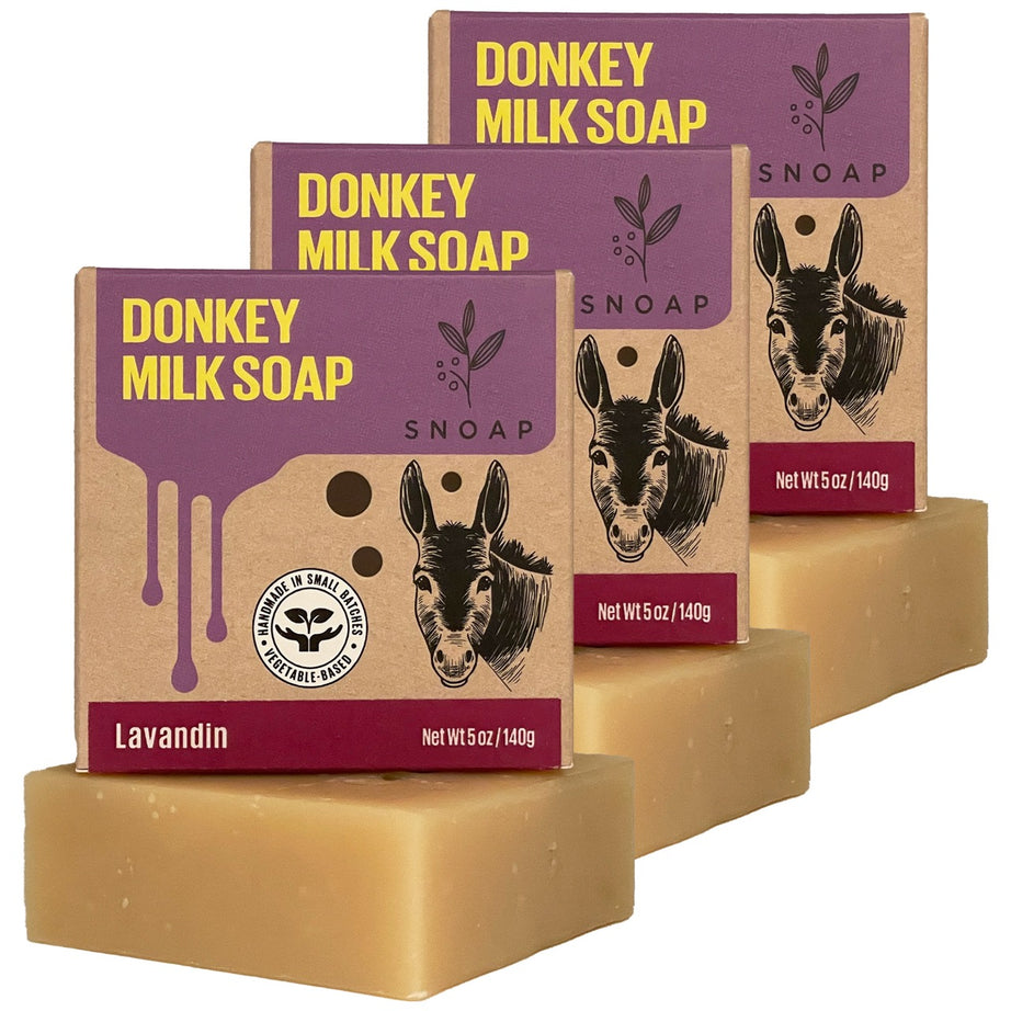 Three bars of lavandin donkey milk soap with a light tan color are stacked in a staggered formation, each below its cardboard box packaging. The boxes are decorated with a sketch of a donkey and the text 'Donkey Milk Soap' and 'SNOAP'.