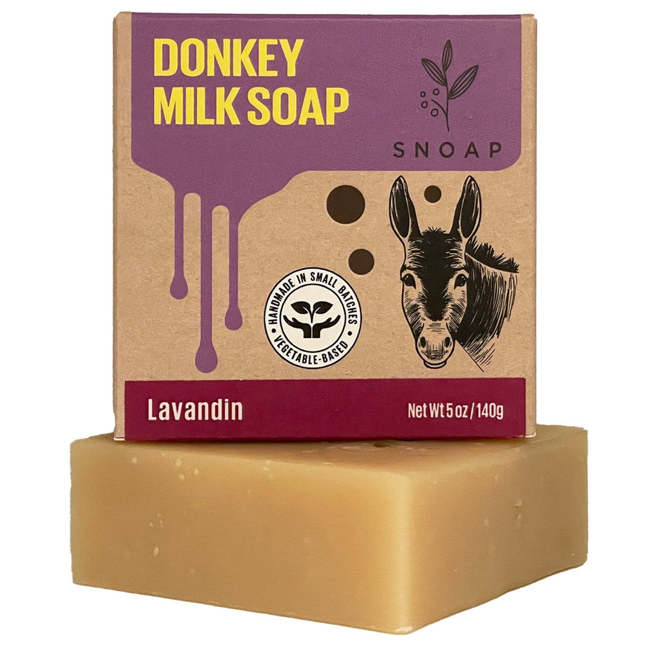 A bar of lavandin donkey milk soap with a light tan color is stacked below its cardboard box packaging. The box is decorated with a sketch of a donkey and the text 'Donkey Milk Soap' and 'SNOAP'.