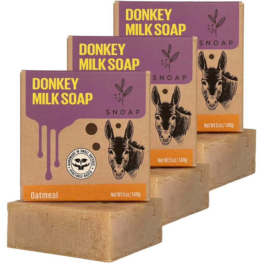Three bars of oatmeal donkey milk soap with a light brown color are stacked in a staggered formation, each below its cardboard box packaging. The boxes are decorated with a sketch of a donkey and the text 'Donkey Milk Soap' and 'SNOAP'.