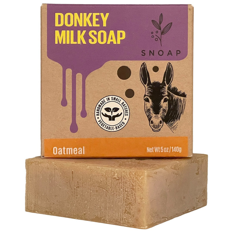 A bar of oatmeal donkey milk soap with a light brown color is stacked below its cardboard box packaging. The box is decorated with a sketch of a donkey and the text 'Donkey Milk Soap' and 'SNOAP'.