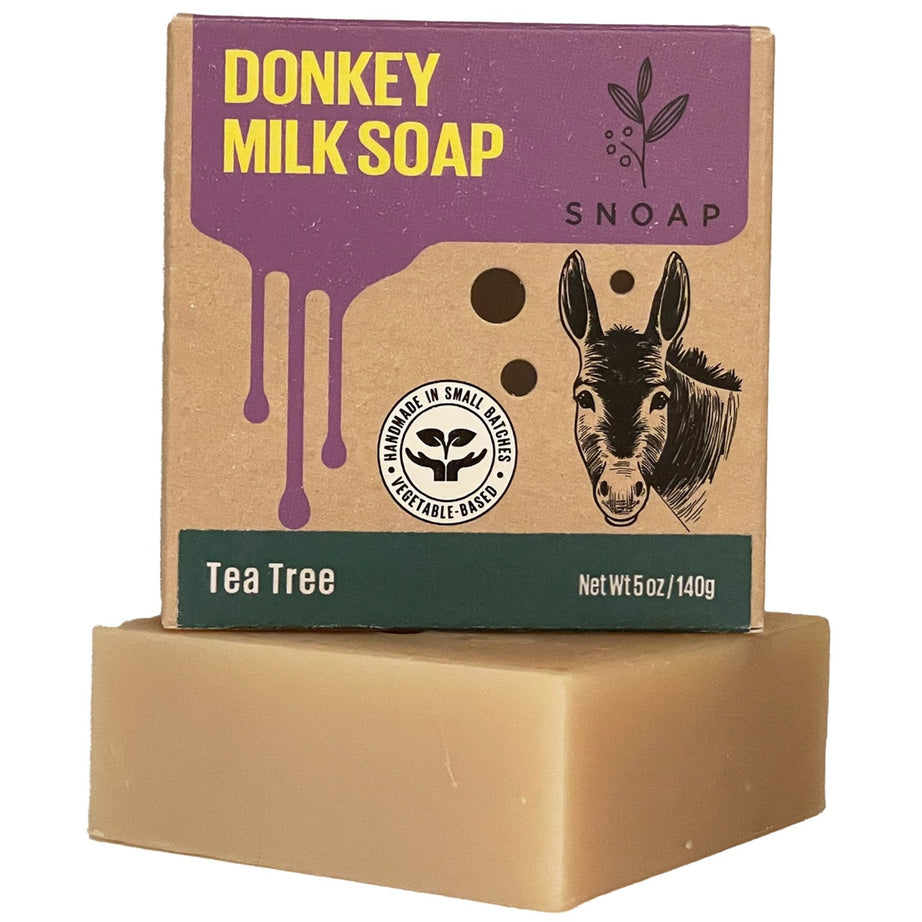 A bar of tea tree donkey milk soap with a light tan color is stacked below its cardboard box packaging. The box is decorated with a sketch of a donkey and the text 'Donkey Milk Soap' and 'SNOAP'.