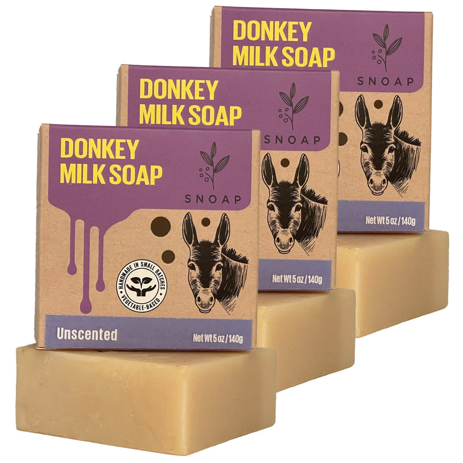 Three bars of unscented donkey milk soap with a light tan color are stacked in a staggered formation, each below its cardboard box packaging. The boxes are decorated with a sketch of a donkey and the text 'Donkey Milk Soap' and 'SNOAP'.
